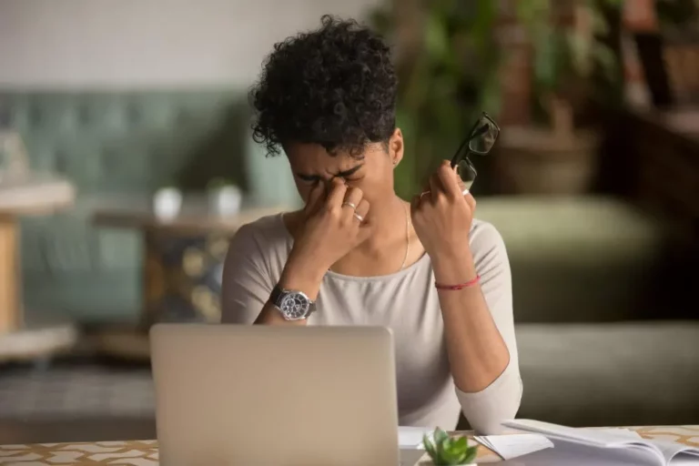 Junge Frau sitzt vor dem Laptop, hält eine Brille in der Hand und verzieht das Gesicht, eine Hand kneift in den Nasenrücken.